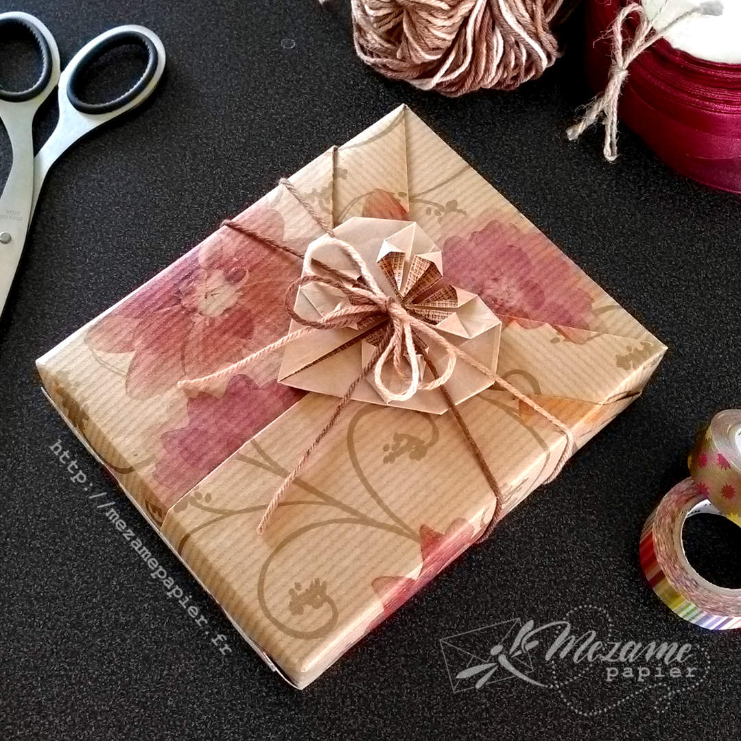 Cadeau emballé dans du papier kraft blond avec impressions de gerbera oranges, avec des plis formant un col de kimono et une décoration origami en forme de cœur