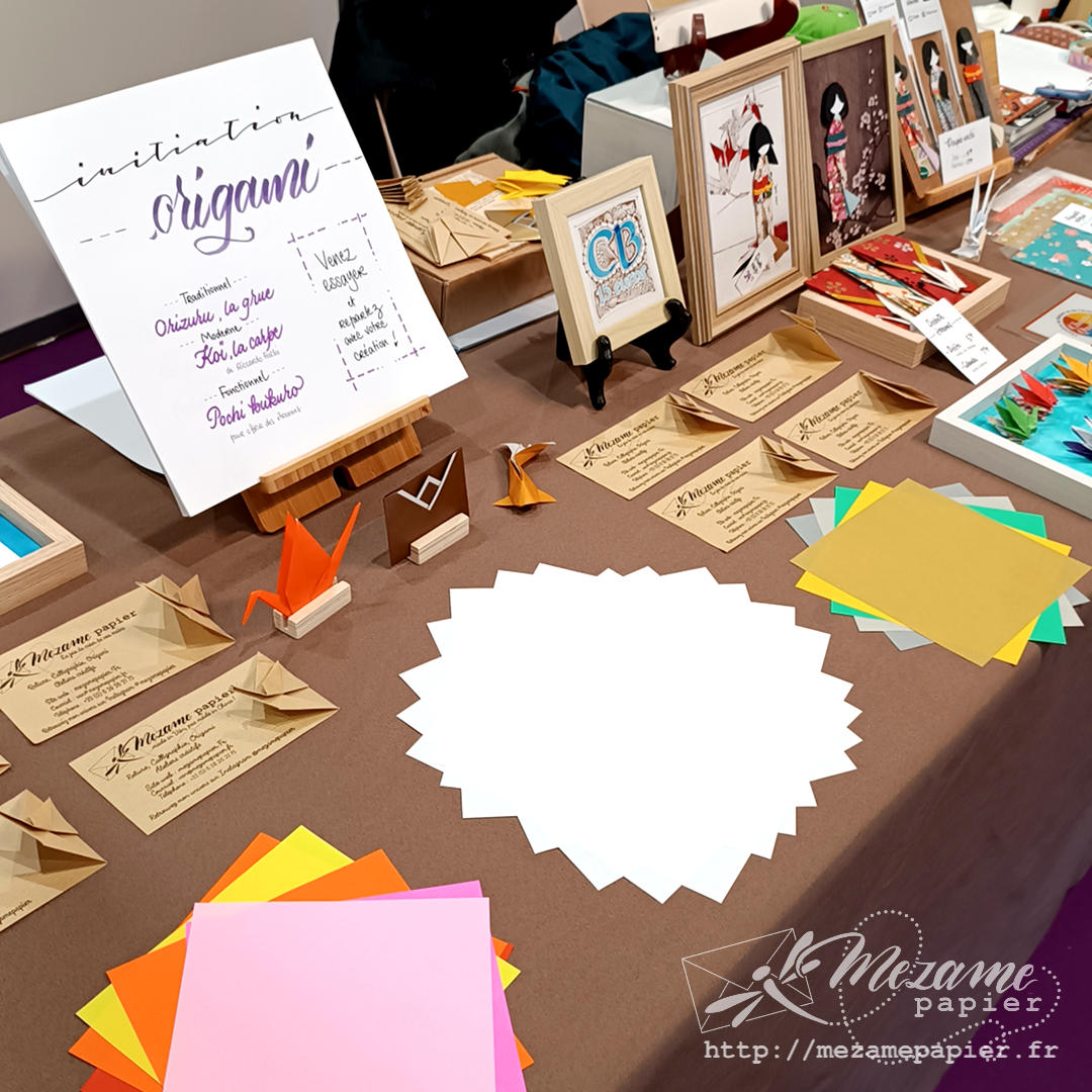 Table avec nappe brune et carrés de papier origami colorés. Panneau écrit à la main annonçant une prochaine initiation origami